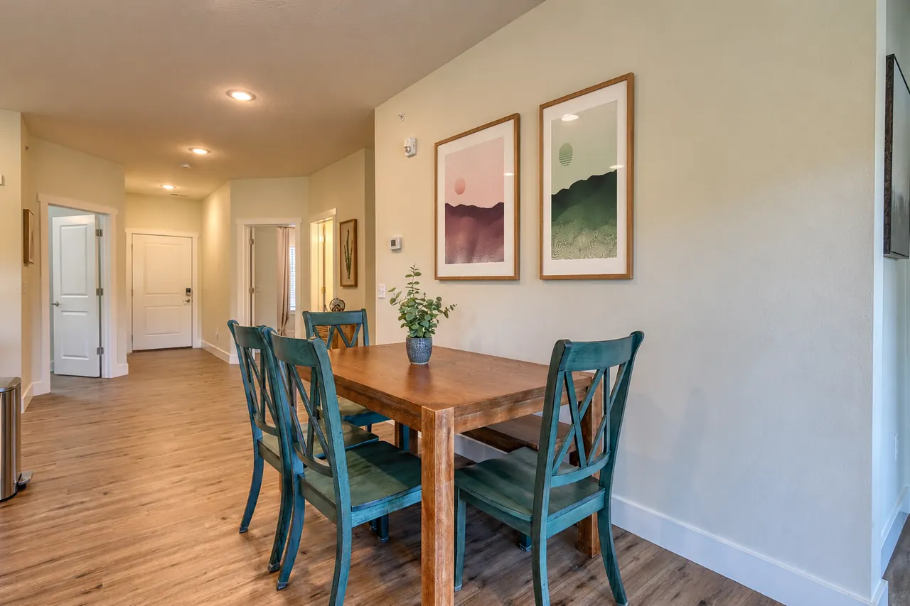 Dining area in a rental apartment with a wooden table, four teal cross-back chairs and a small potted plant. On the pale green wall above hang two framed Ridgeline prints in oak frames: on the left, blush-pink hills under a pale sun; on the right, deep green striped moorland under a green sun. A hallway with white doors and wood-look floors leads off to the left.